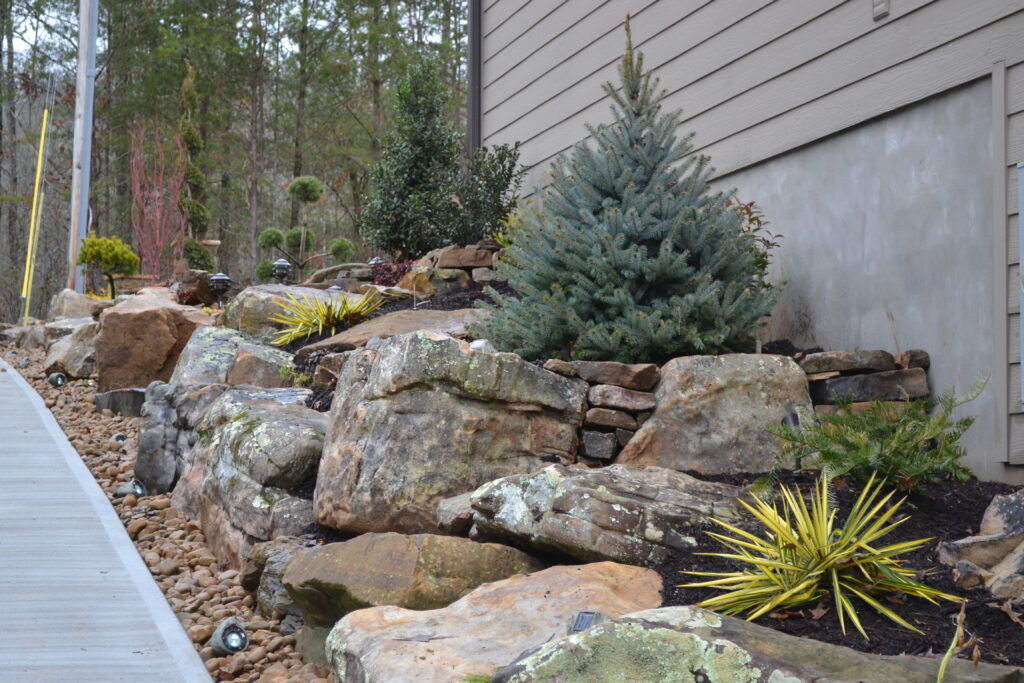 Outdoor Space - Boulder retaining wall with evergreen plantings and decorative rock landscaping design. - Garden Guru Services