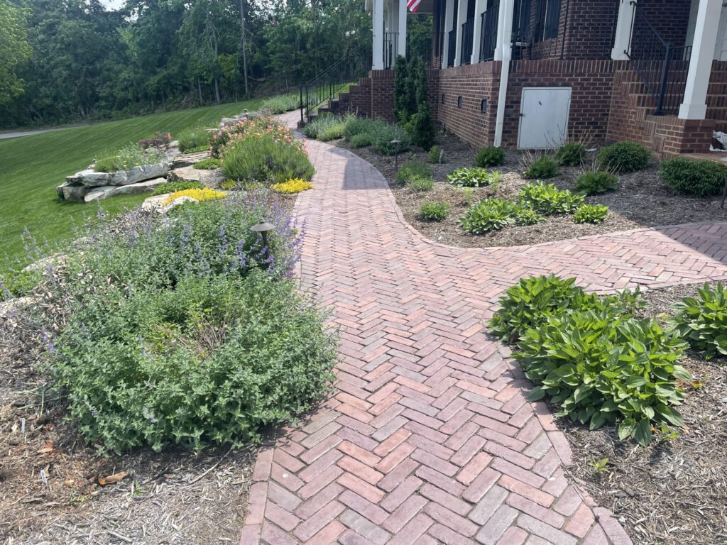 Outdoor Space - Curved brick walkway with herringbone pattern, perennial garden beds, and foundation plantings for front yard landscape design. - Garden Guru Services