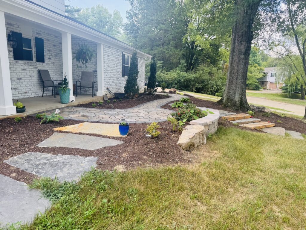 Outdoor Space - Front yard landscape renovation with stone patio, natural rock retaining wall, mulched garden beds, and mature tree preservation. - Garden Guru Services